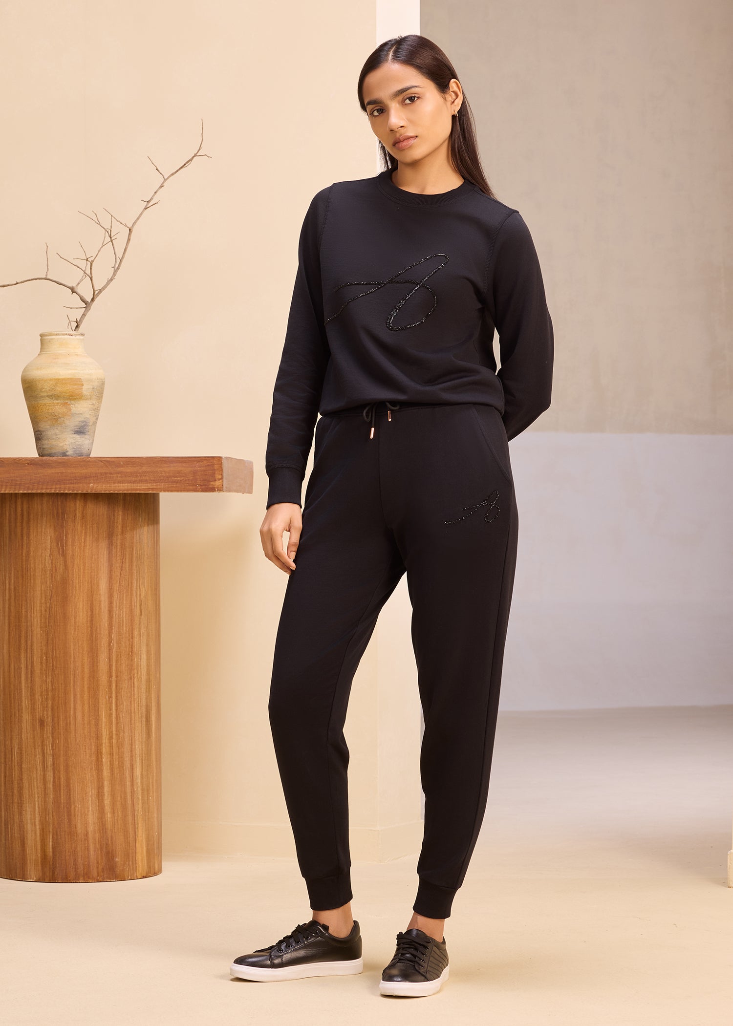 Female model wearing a black sweatshirt and joggers with matching black sneakers, standing in a minimalistic setting with neutral tones and a wooden table with a decorative vase.