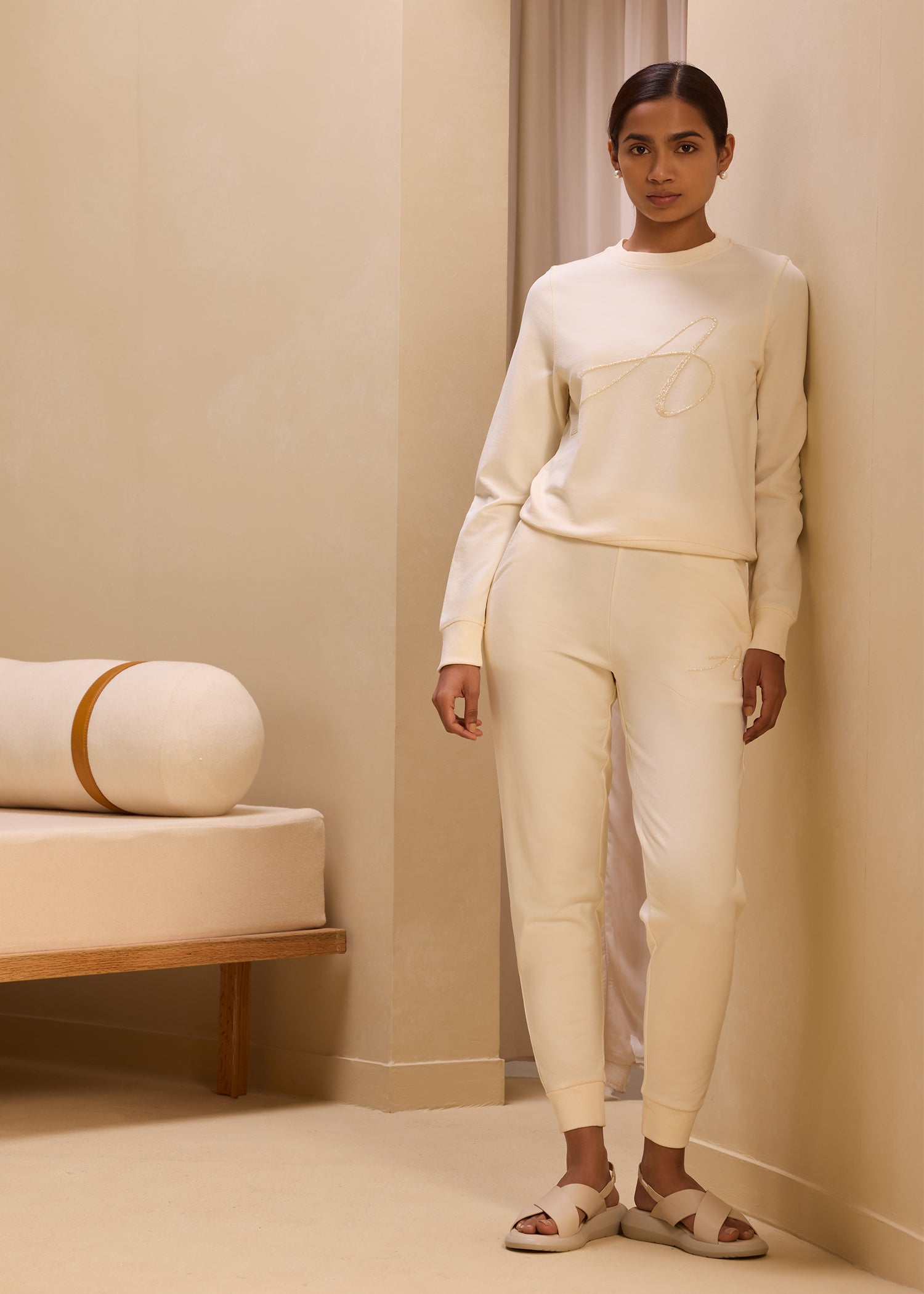 Model wearing a matching cream-colored jogger set with embroidered 'A' logo on the sweatshirt and pants, exuding a minimalist and cozy aesthetic in a neutral-toned room.