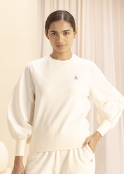 A woman wearing a minimalist, light-colored sweatshirt and matching pants with a delicate embroidered emblem, standing indoors against a neutral backdrop.