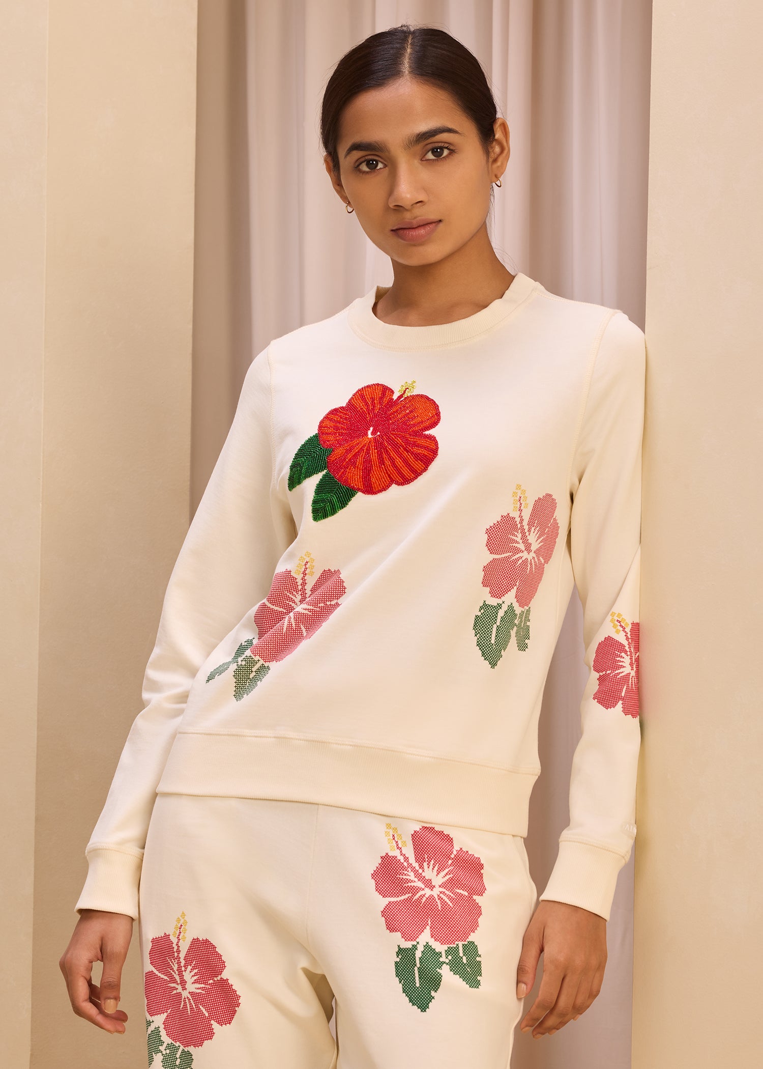 Model wearing a cream-colored sweatshirt with vibrant embroidered hibiscus flower design and matching pants, set against a neutral background