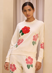 Model wearing a cream-colored sweatshirt with vibrant embroidered hibiscus flower design and matching pants, set against a neutral background