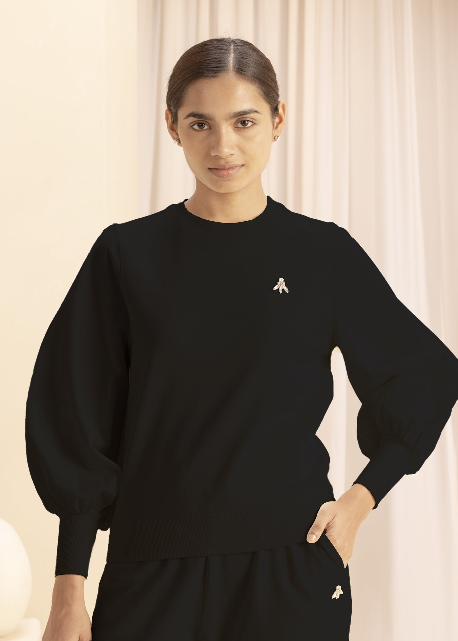 Young woman stands indoors wearing a black sweatshirt with puffed sleeves and a small emblem on the chest. 