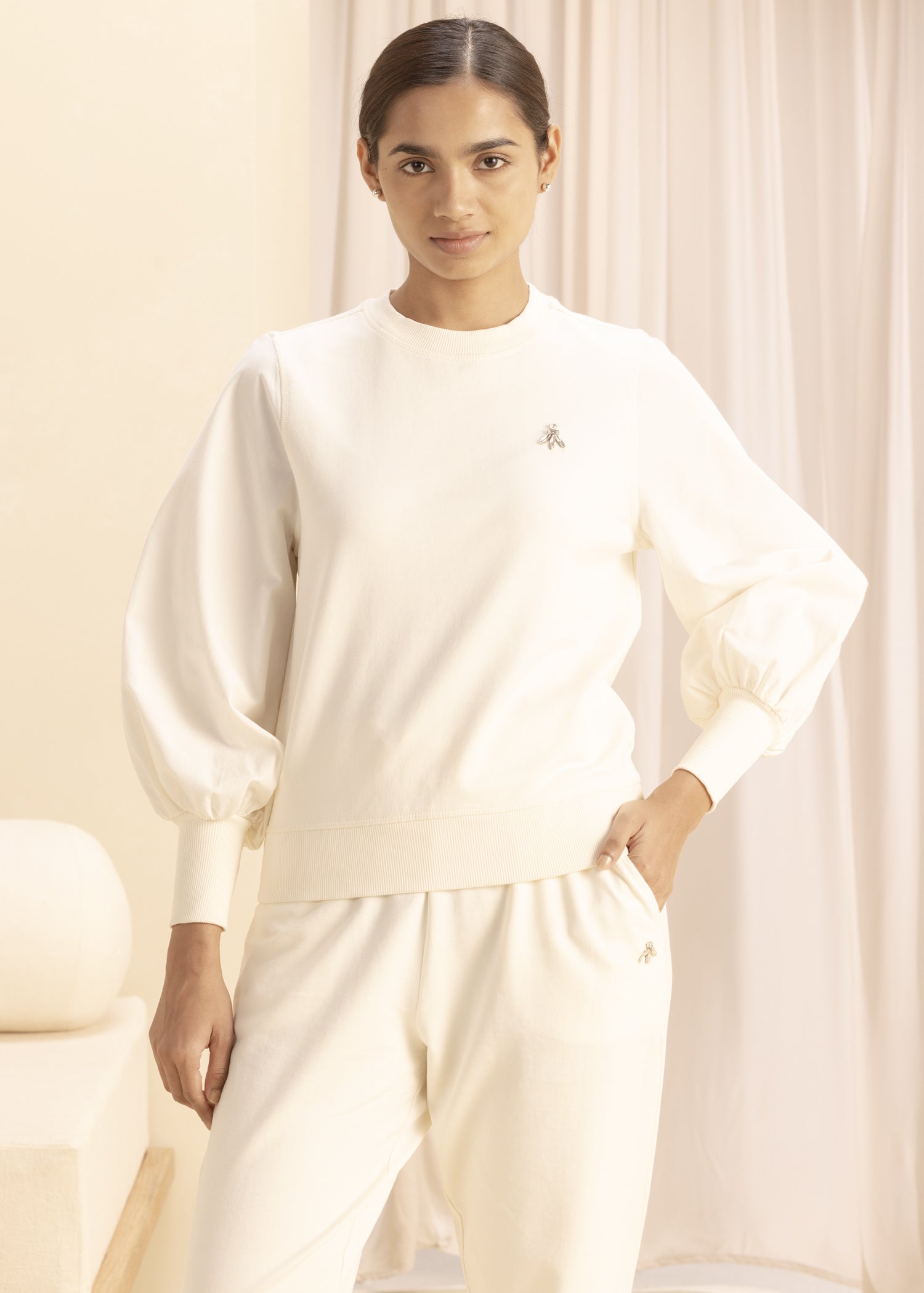 Model wearing cream-colored sweatshirt with puffed sleeves, featuring a minimalist design and comfortable fabric, standing in a softly-lit room with neutral decor.