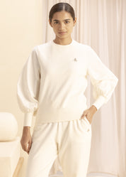 Model wearing cream-colored sweatshirt with puffed sleeves, featuring a minimalist design and comfortable fabric, standing in a softly-lit room with neutral decor.